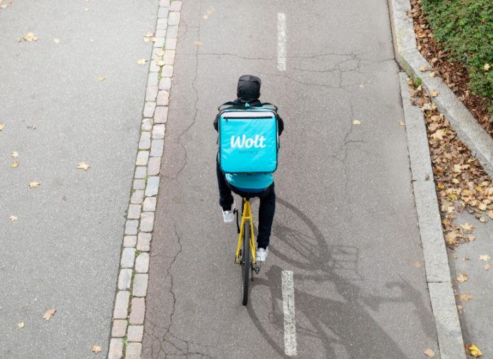 Aerial view of a bicycle courier on an open road wearing a large blue Wolt-branded backpack.