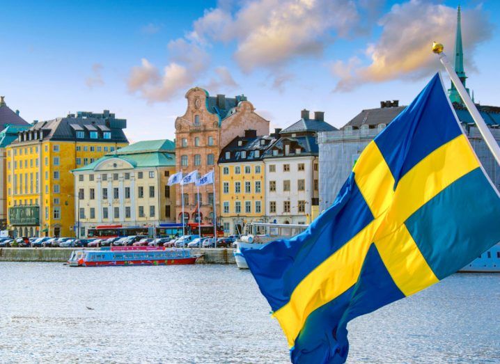 Swedish flag waving in the foreground of a shot of a Swedish cityscape in the background.