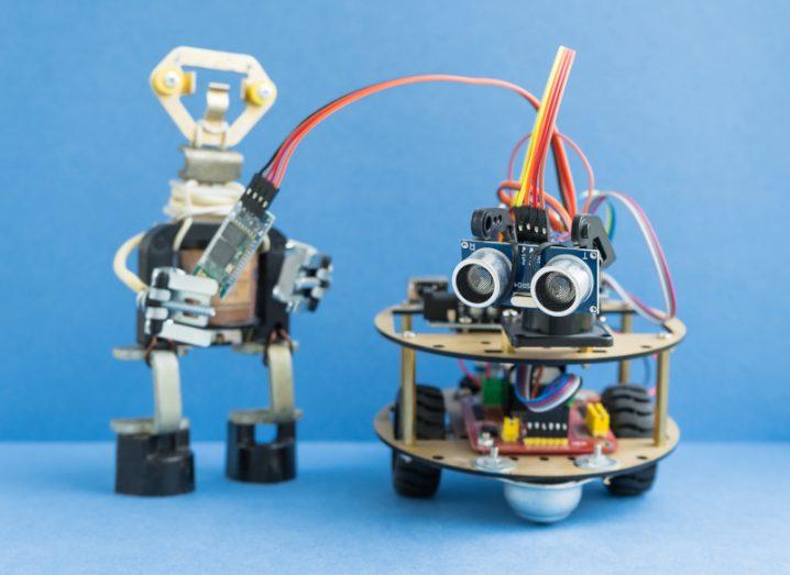 A small homemade robot shaped like a stick figure holds a wired component of a second robot next to it, with exposed electronics and small lightbulbs for eyes.