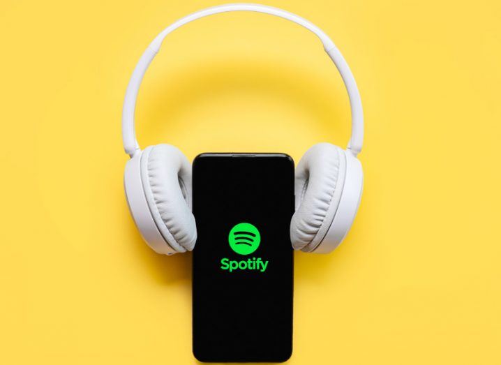 White headphones curled around a phone with the Spotify logo on its screen against a bright yellow background.