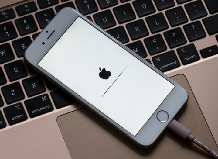 An iPhone is plugged in, sitting on top of a keyboard. The screen is showing a loading page with the Apple logo on it.