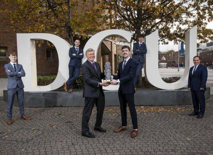 DCU student receives trophy from ESB's representative as his three teammates and coach watch on. They are standing at a huge DCU sign made up of the letters in white.