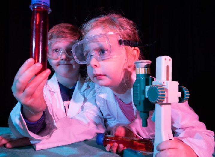 A child looking intently at a test tube while another child stands behind her looking at it. Both are wearing protective lab gear.