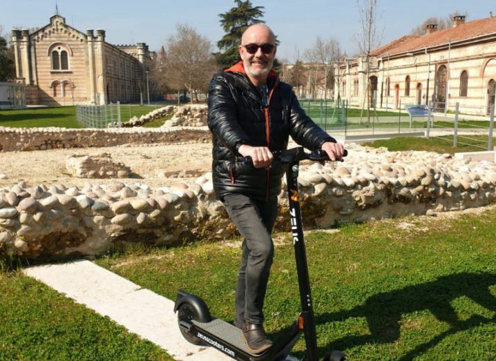Damian Young of Zeus riding an e-scooter in a garden on a sunny day.