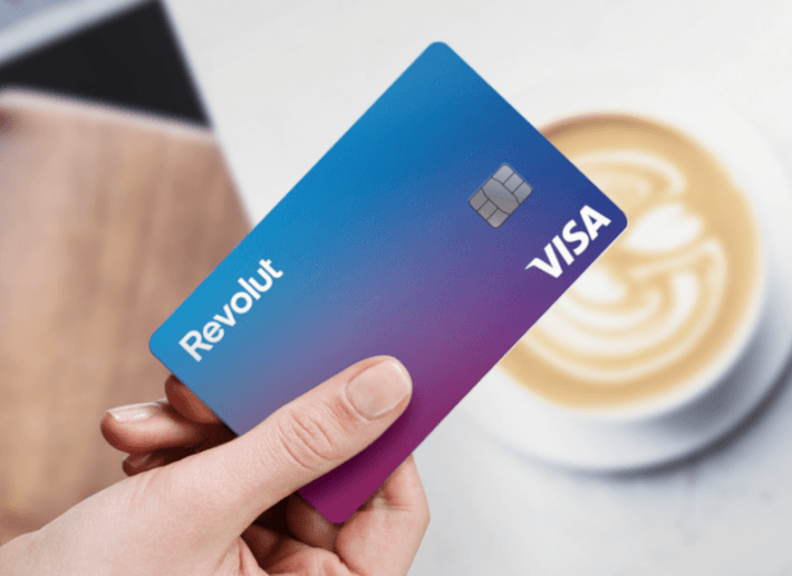 A person’s hand is holding a Revolut card above a table in a cafe, with a cup of coffee in the background.
