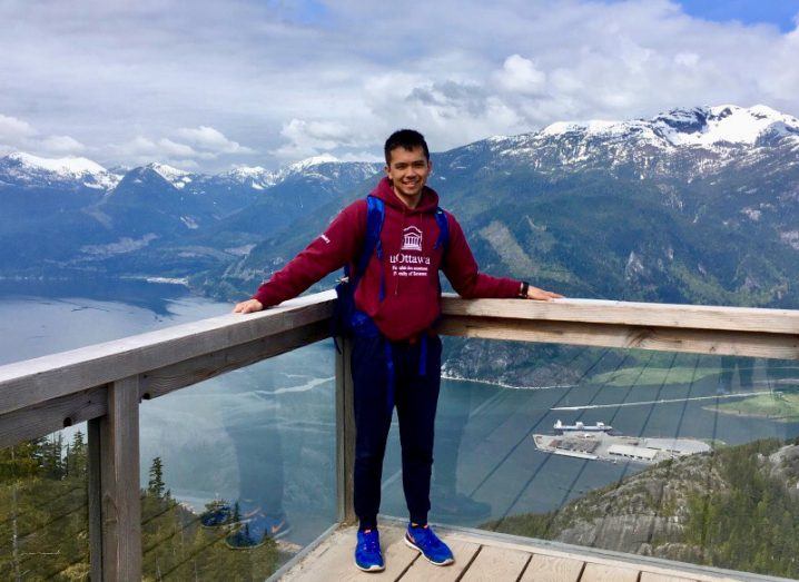 Kevin Mercurio stands on a balcony overlooking a lake at the foot of a large snow-capped mountain range. He is wearing a hoody and a backpack.