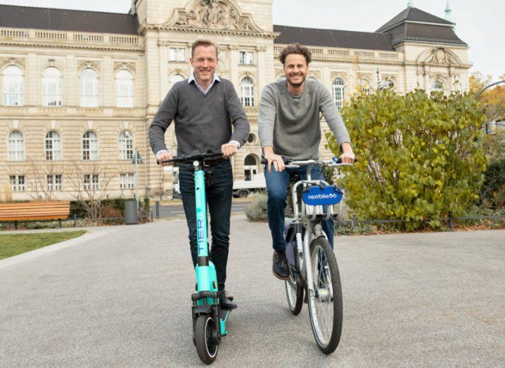 Leonhard von Harrach and Lawrence Leuschner on a Tier scooter and a Nextbike bicycle in the garden of a grand sandstone building.