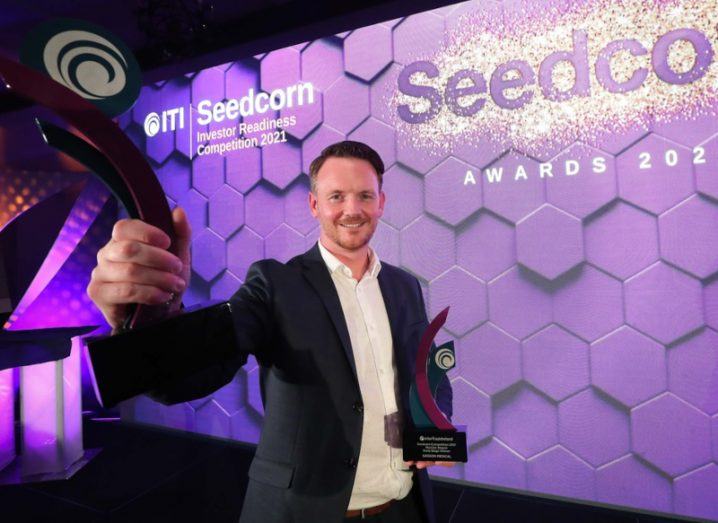 A man in a suit holding two statues in front of a purple wall with the Seedcorn logo on it.