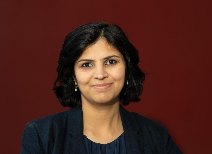 A woman with short, dark hair wearing a navy business jacket against a dark red background. She’s smiling at the camera.