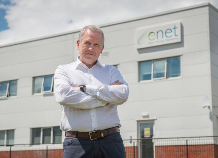 A man stands with his arms crossed outside a large building that says 'Enet' on the side of it.