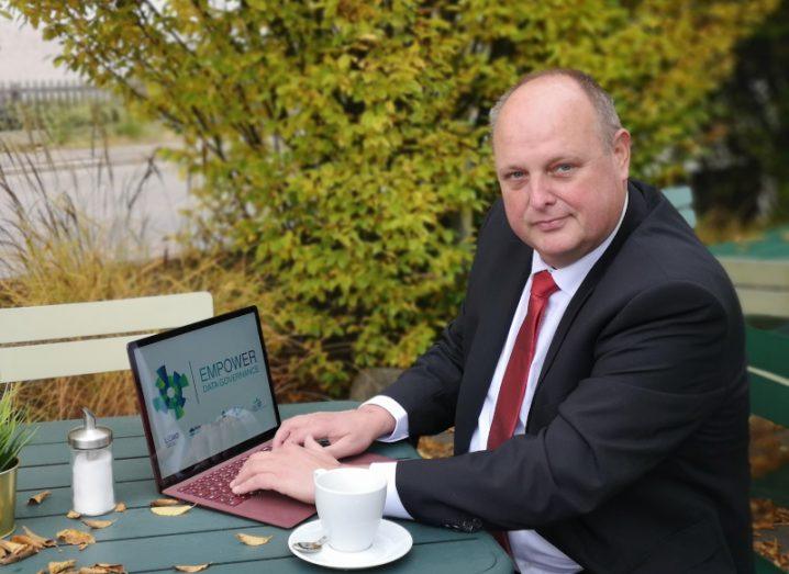 A man in a suit sitting at an outdoor table with a coffee and a laptop. The screen on the laptop says Empower.