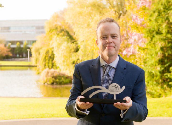 A man in a suit stands near the lake on the UCD campus, holding up a Science Foundation Ireland Award.