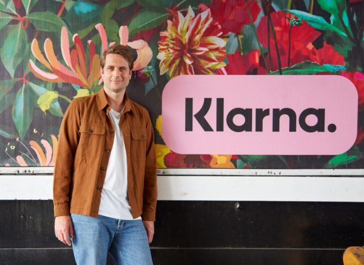 Klarna CEO Sebastian Siemiatkowski stands against a wall decorated with bold floral wallpaper and the blush pink Klarna logo.