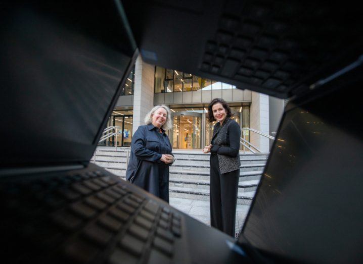 Two women stand outside the steps of a large building, framed by a two open laptops forming a square shape around their figures.