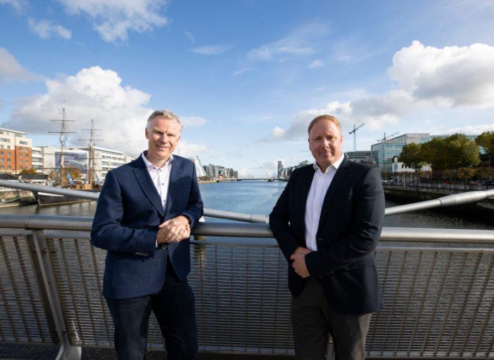 ActionPoint CEO David Jeffreys and ICT founder Barry Byrne standing on a Dublin bridge over the Liffey. Dublin docklands and Samuel Beckett bridge can be seen in the distance.