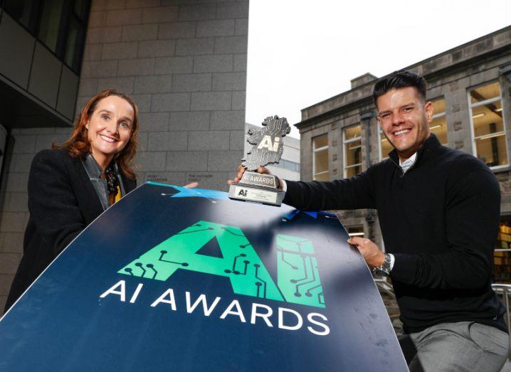 A woman and a man stand apart while holding a sign that says AI awards in large writing. The man is holding a small award trophy.
