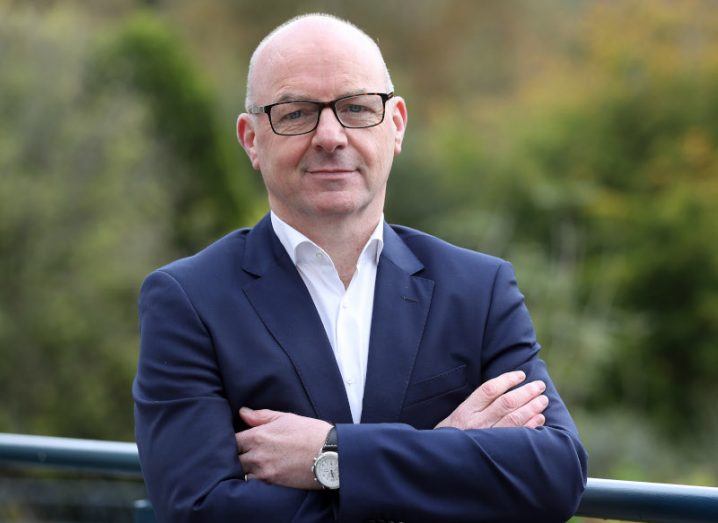 Lorcan Malone, CEO of IDA Ireland, posing for a photo with arms folded. He is wearing a navy blue suit and black glasses. Blurred trees in the background.