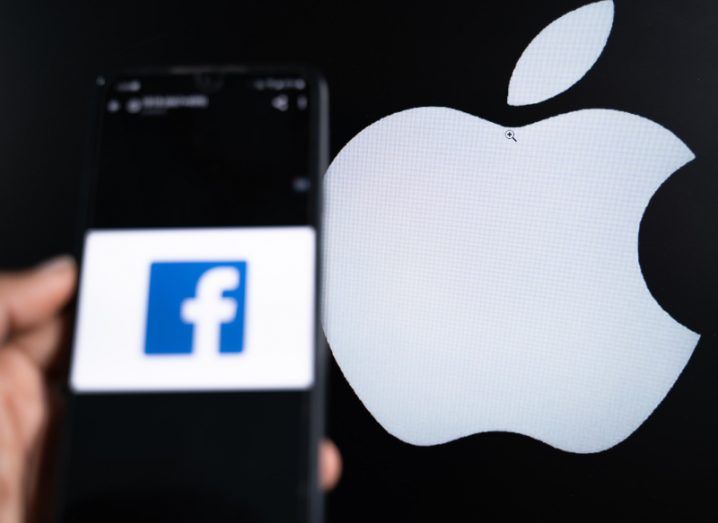 Facebook logo on smartphone screen held in someone's hand. The Apple logo can be seen in the background.
