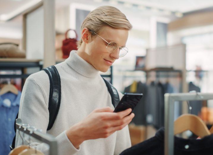 Man wearing glasses and a white turtleneck sweater with bag behind his back holds a smartphone while shopping in a retail store.