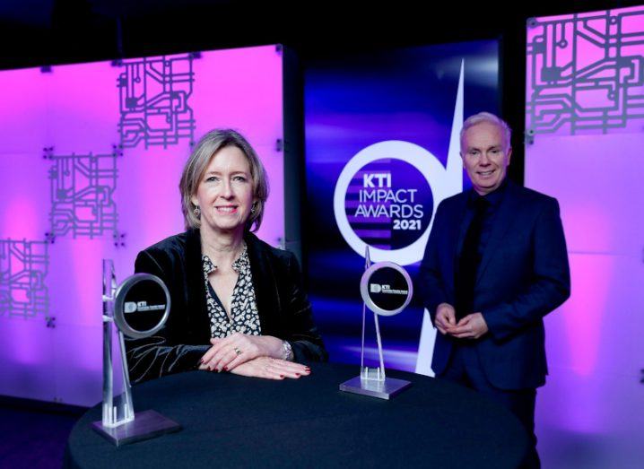 KTI Director Alison Campbell and Impact Awards MC Richard Curran standing behind a table with the award trophies placed on it. KTI Impact Awards branding is on a poster in the background.