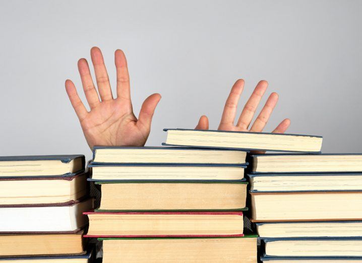 A pair of hands raised behind a stack of books in front of a grey backgorund.