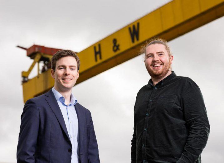 Two men pictured against an overcast sky with the iconic Belfast landmark of the yellow Harland & Wolff crane visible in the background.