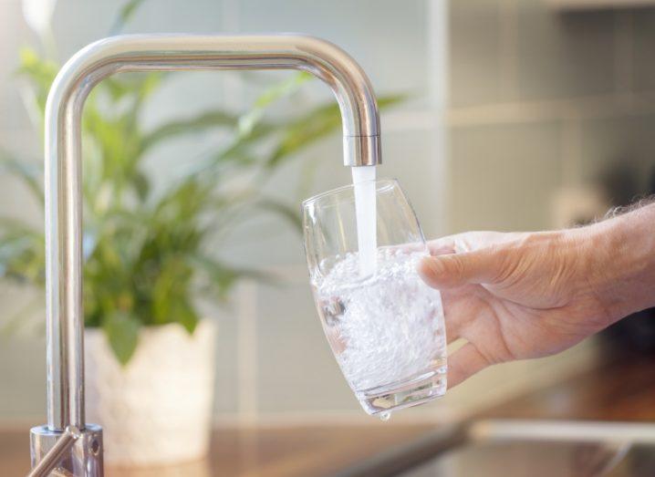 Image of a man's hand holding a glass in front of a kitchen tap with water pouring out.