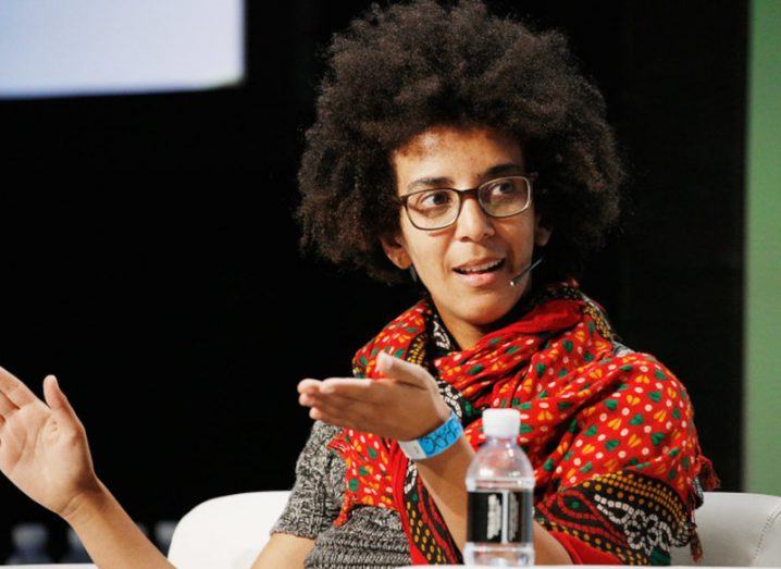 Timnit Gebru speaking at a TechCrunch event with a water bottle in front of her and the dark background of the stage behind her.