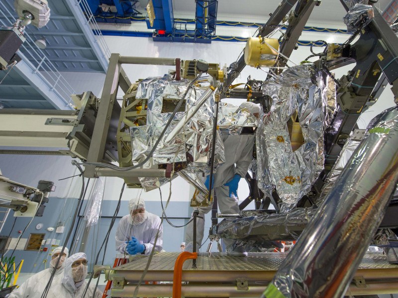 People in protective gear are working on a large spacecraft instrument.