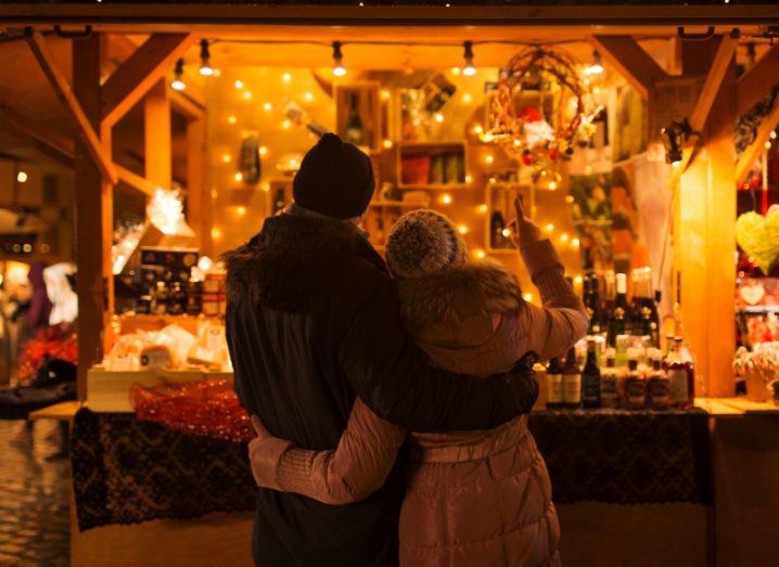 Man and woman standing in front of a christmas market in the evening time with lights around the stall.
