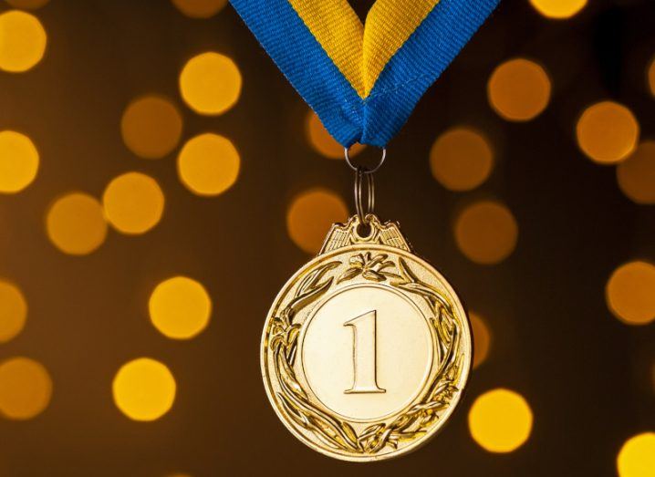 Gold medal with number one on it hanging from a blue and yellow striped ribbon against glittering gold background.