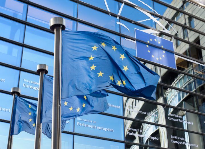 European Union flags in front of a large windowed building, with the reflections showing a clear blue sky.
