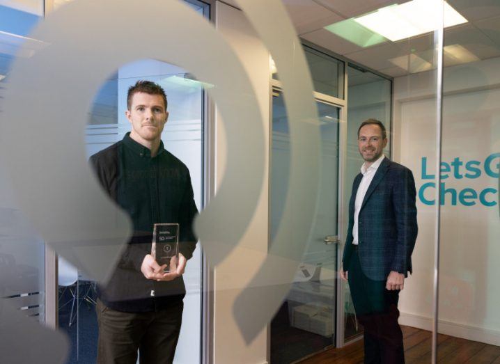 Two men stand in a room with 'LetsGetChecked' written on the window. One of the men is holding an award.