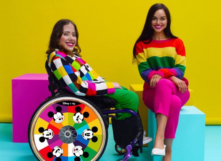 A young woman sits in a brightly coloured wheelchair, beside another young woman in a bright outfit.