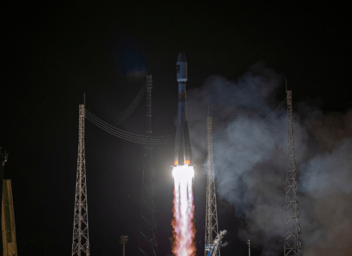 A rocket taking off from a spaceport, the engines are lit as the rocket leaves a trail of flame and smoke behind it with a night sky background.