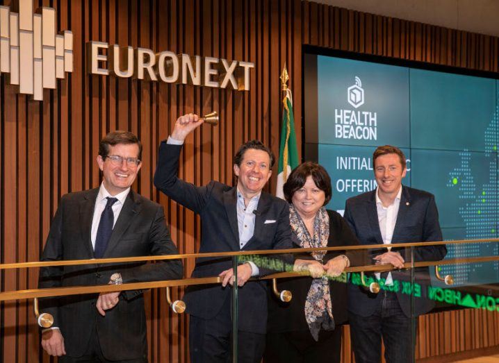 Three men and a woman stand at a railing with the Euronext logo on the wall behind them along with a screen that says HealthBeacon. One man is ringing a small bell.