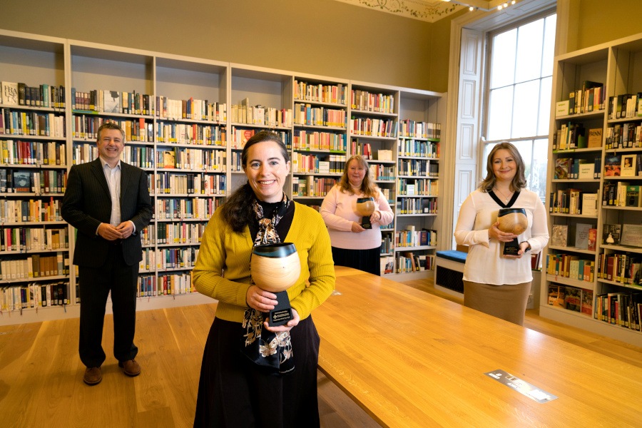 A man stands in a library with three women, each of whom are holding an award.