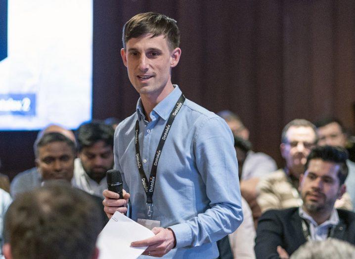 A man standing at a conference in the centre of a seated crowd. He is holding a microphone and wearing a blue shirt and lanyard.