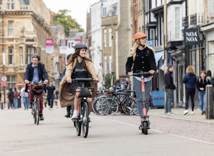 Three people use Voi e-scooters on the streets of Cambridge surrounded by people and old, stone buildings.