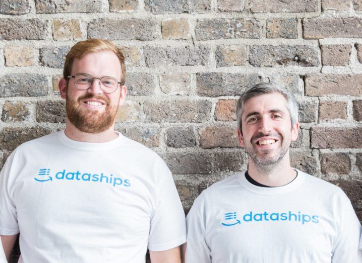 Two men wearing white T-shirts bearing the Dataships logo standing against an exposed brick wall.