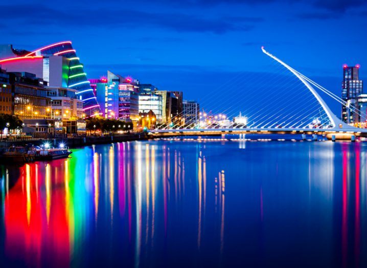 Skyline of Dublin's port and financial district. Multi-coloured lights from buildings reflecting on the Liffey river, with the Samuel Beckett bridge in the distance.