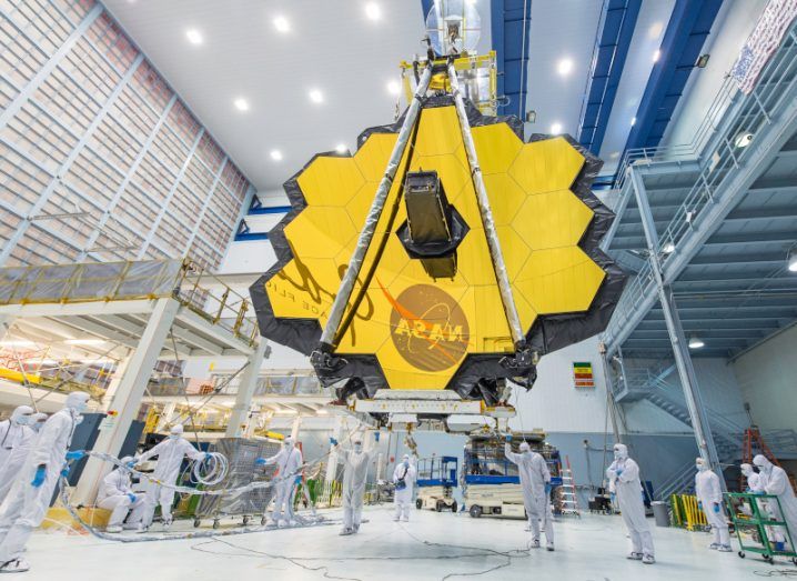 The James Webb telescope’s 18-segmented gold mirror section seen in full bloom while being lifted by a crane in an indoor NASA centre with scientists wearing white industrial suits standing around it. The NASA logo is reflected on the lower section of the mirror.