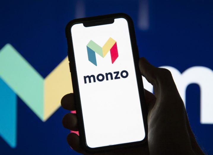 Monzo logo on a smartphone screen held in a person's hand. Larger Monzo logo in the background.