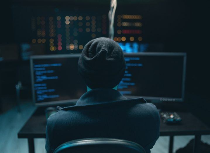Hooded hacker sitting in front of two computer screens in a dark room.