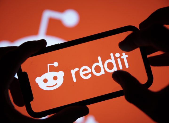 Silhouette of hands holding a smartphone with the reddit logo on the screen, and a blurred reddit logo in the background.
