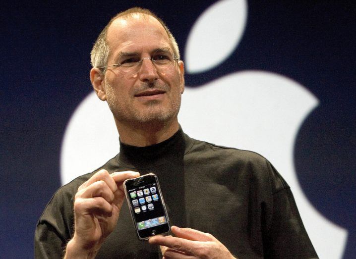 Steve Jobs stands in front of an Apple logo holding an iPhone.