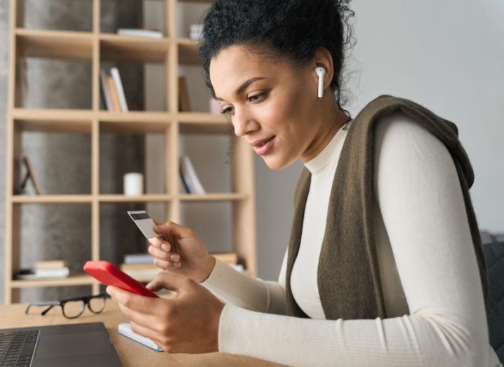 Woman holding a phone in one hand and a credit card in the other. Bookshelf in the background.