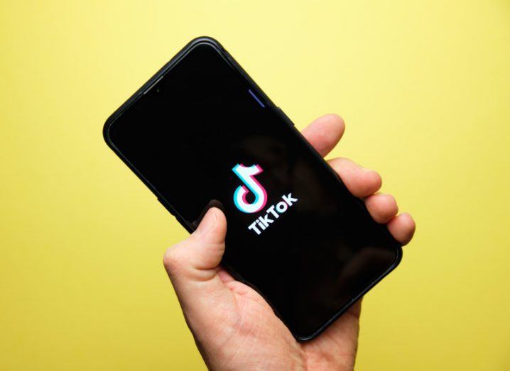 Person's hand holding a smartphone with TikTok logo on screen. Bright yellow background.