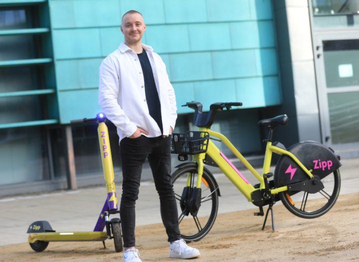 Charlie Gleeson stands outside a building on the UCD campus in front of an e-bike and e-scooter branded with Zipp Mobility’s vibrant colour scheme.