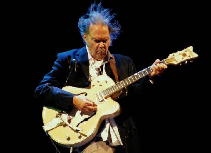 Neil Young holding a guitar performing with a dark background.